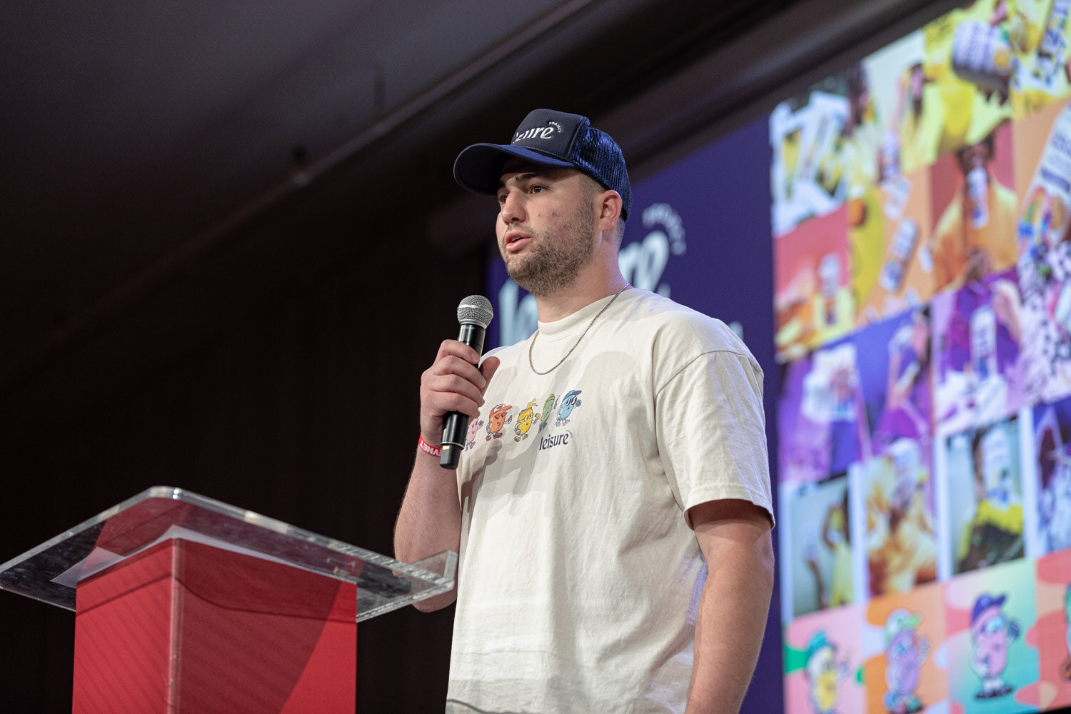 A man in a navy Leisure cap and cream t-shirt holds a microphone beside a podium with a colorful slide behind him.