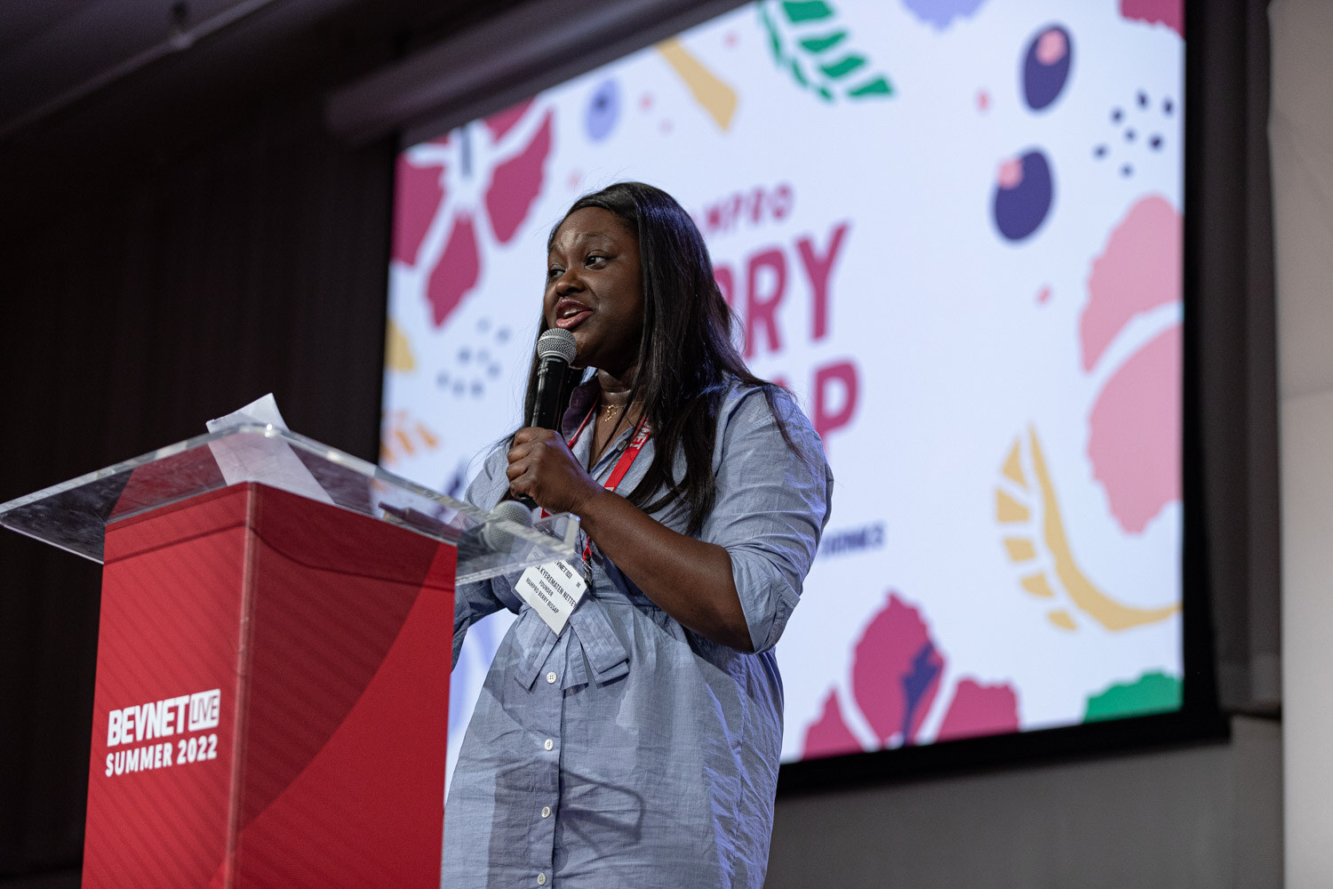 A woman in a blue button-up dress holds a microphone at a red podium in front of a colorful slide reading Berry Bissap.