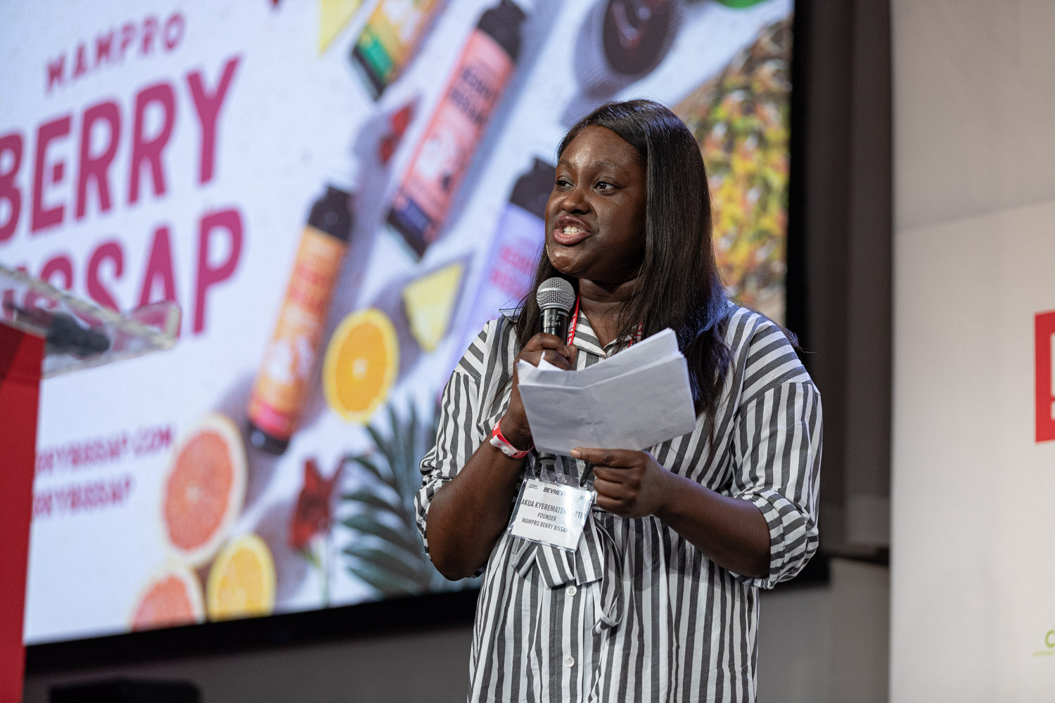 A woman in a black and white striped shirt holds a microphone and papers in front of a slide with Mampro Berry Bissap bottles and citrus fruit.