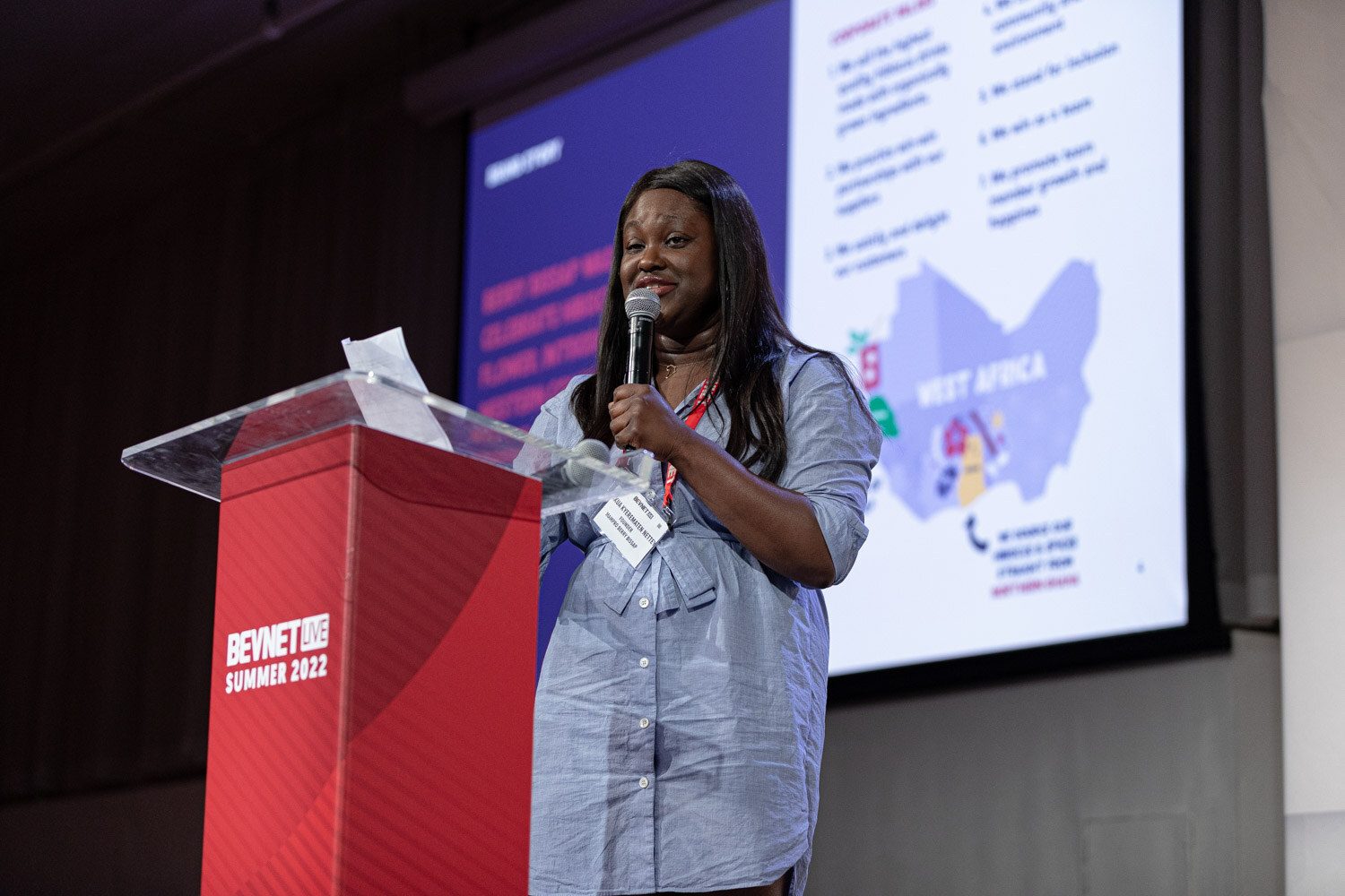 A woman in a blue button-up dress holds a microphone at a red podium in front of a slide with a West Africa map.