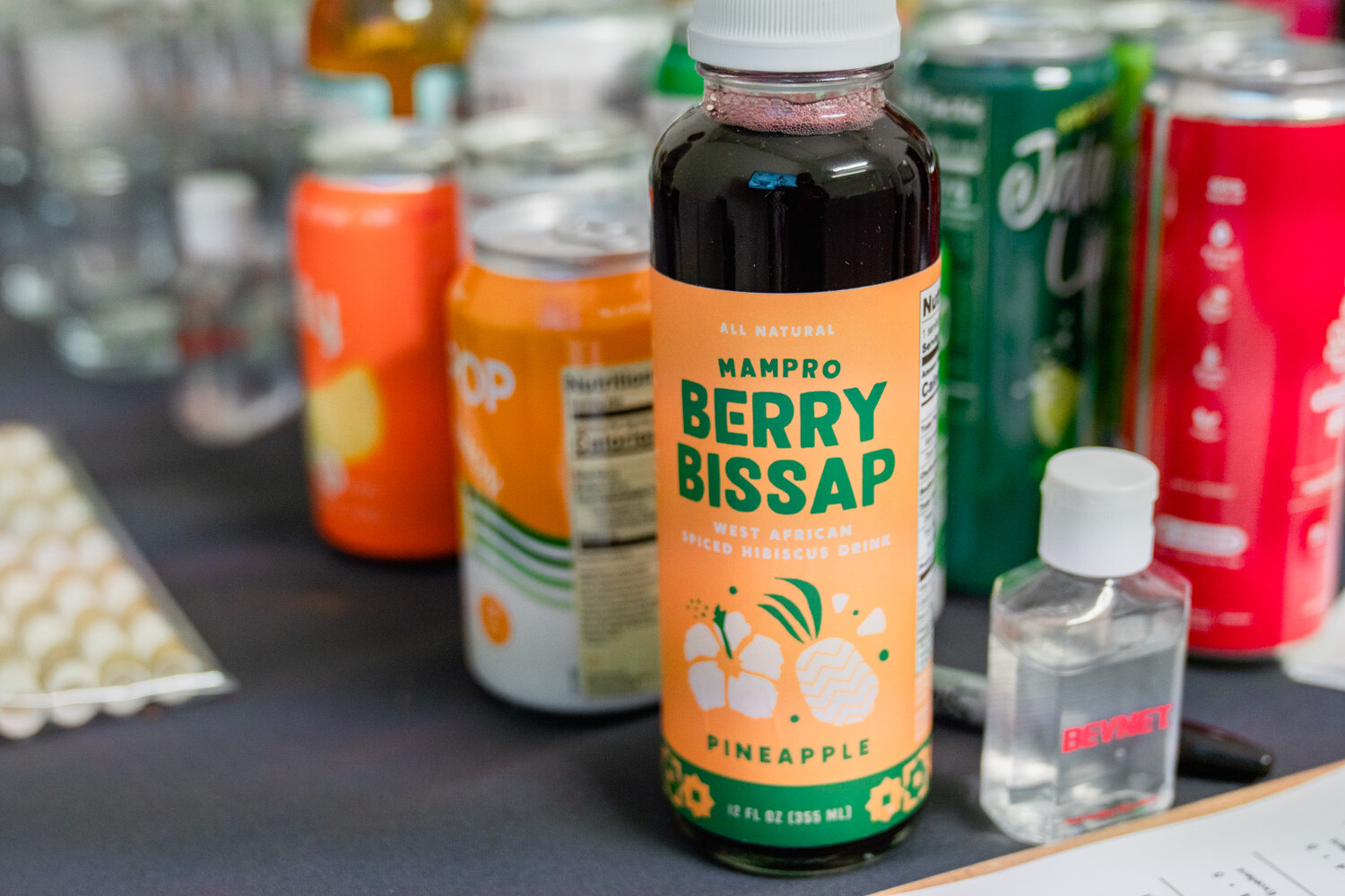 Close-up of a Mampro Berry Bissap pineapple hibiscus bottle with a hand sanitizer bottle and cans of drinks around it.