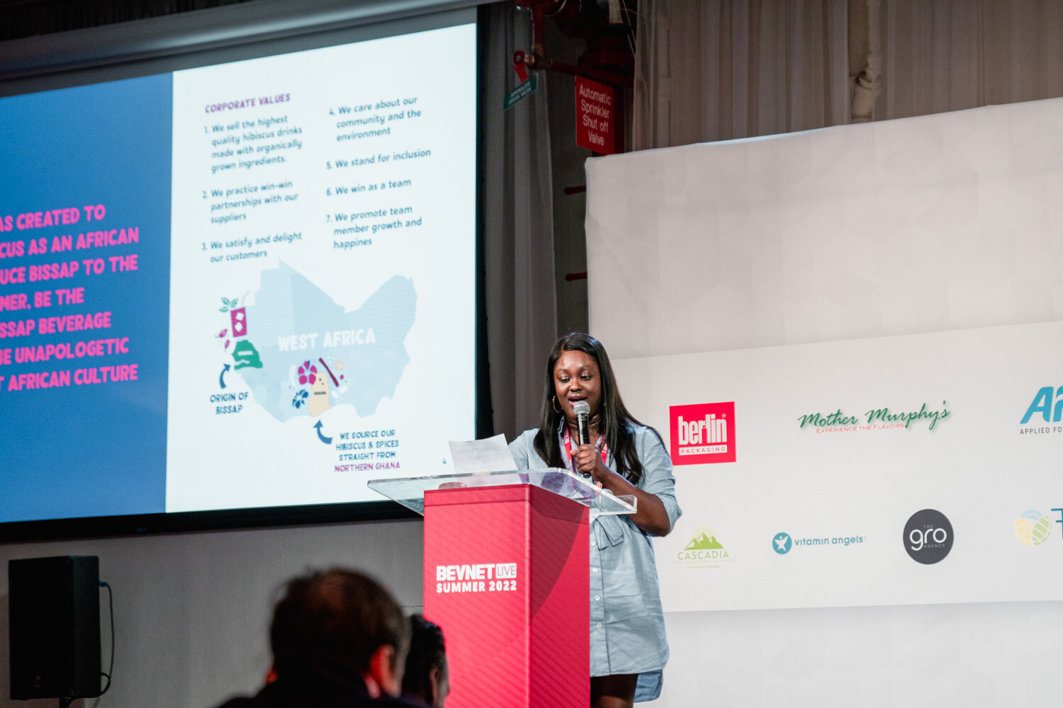 A woman at a red podium holds a microphone in front of a blue and pink slide about corporate values and West Africa.
