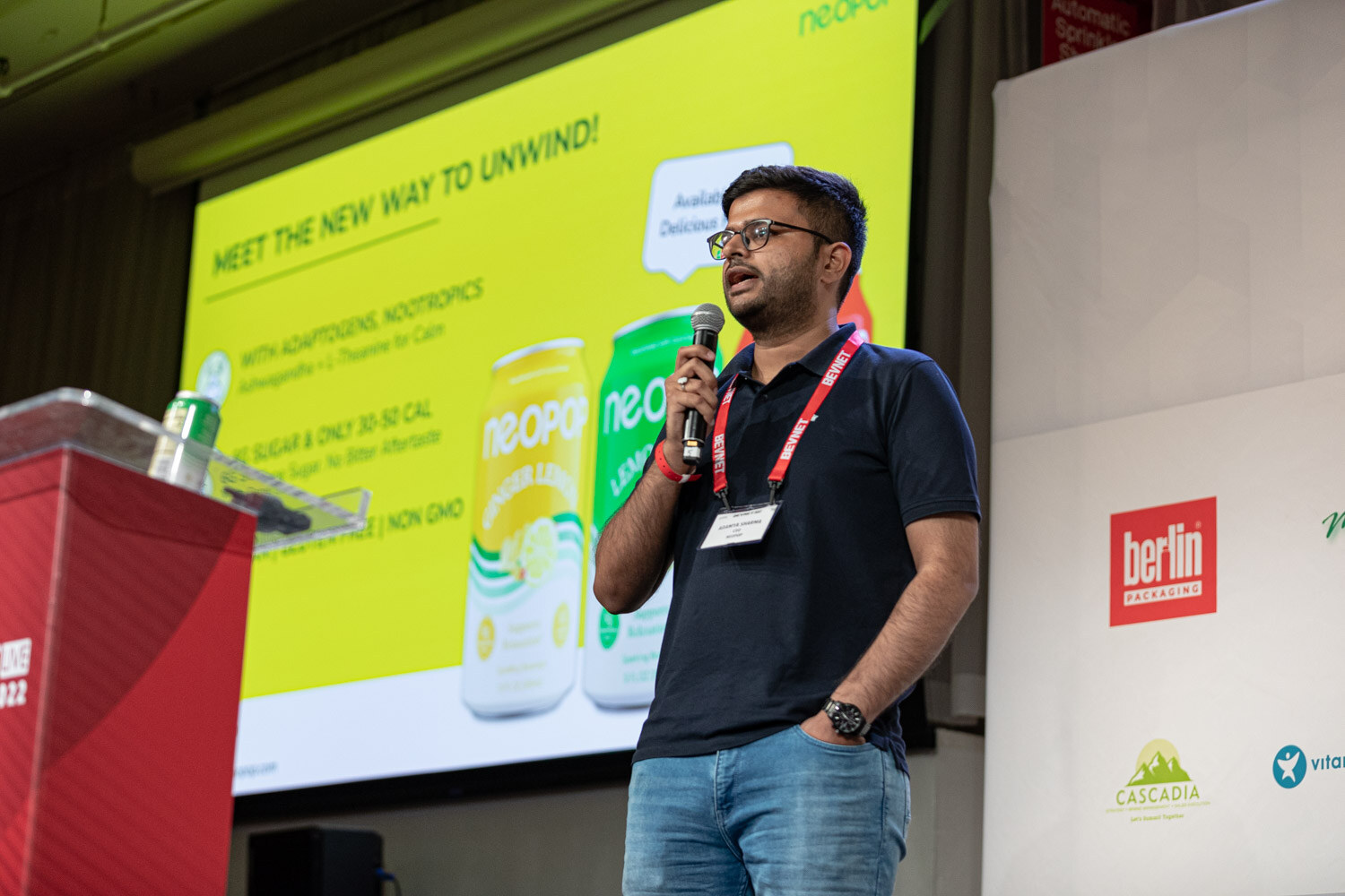 A man in a navy polo with a lanyard holds a microphone in front of a yellow slide of Neopop cans.