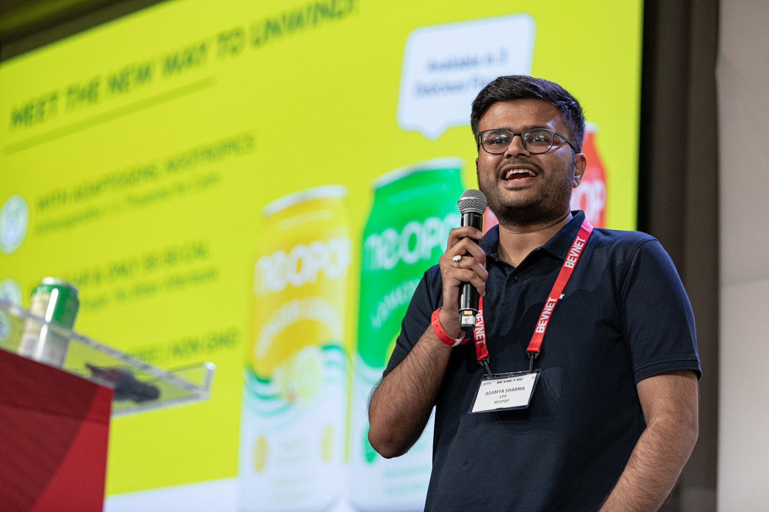 A man in a navy polo and glasses holds a microphone in front of a yellow slide with Neopop Lemon Lime cans.