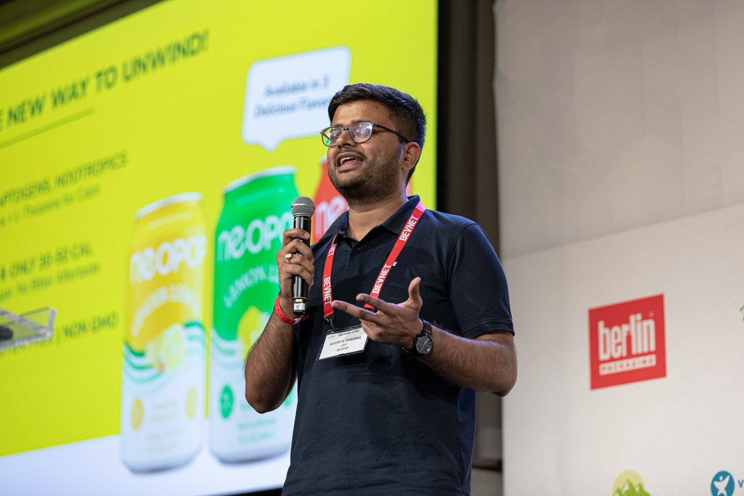 A man in glasses and a navy polo holds a microphone and gestures with one hand in front of a yellow slide showing Neopop cans.