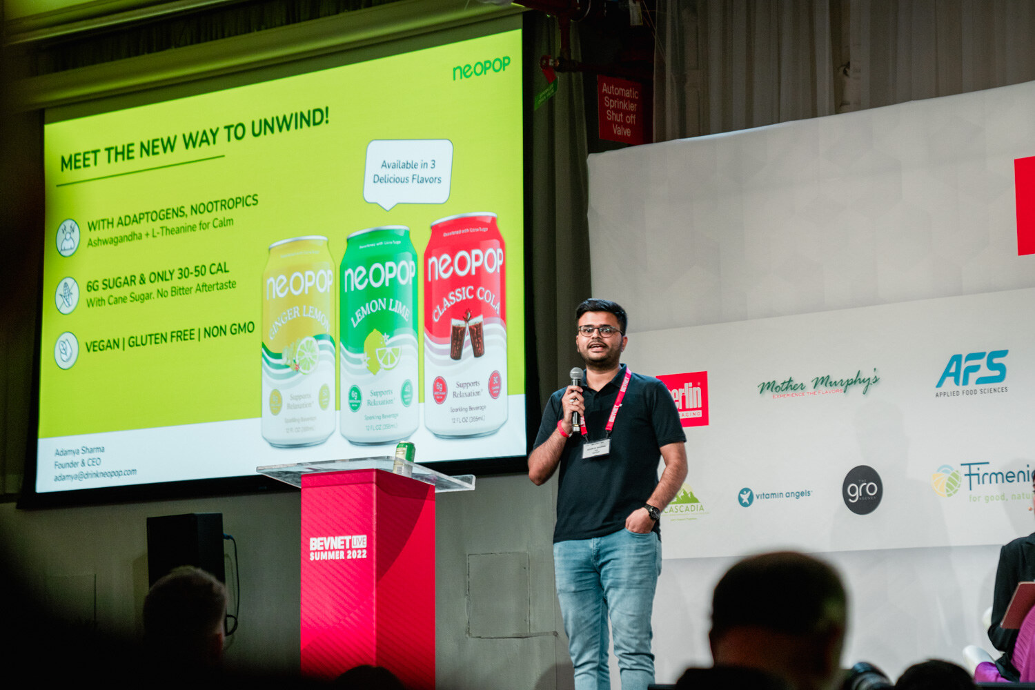 A man in a navy polo and lanyard stands next to a red podium in front of a yellow slide reading Meet the New Way to Unwind with three Neopop cans.