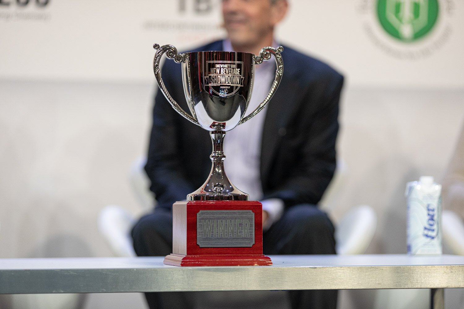 A silver cup labeled New Beverage Showdown Winner on a red base in sharp focus, a blurred man in a navy jacket seated behind it.