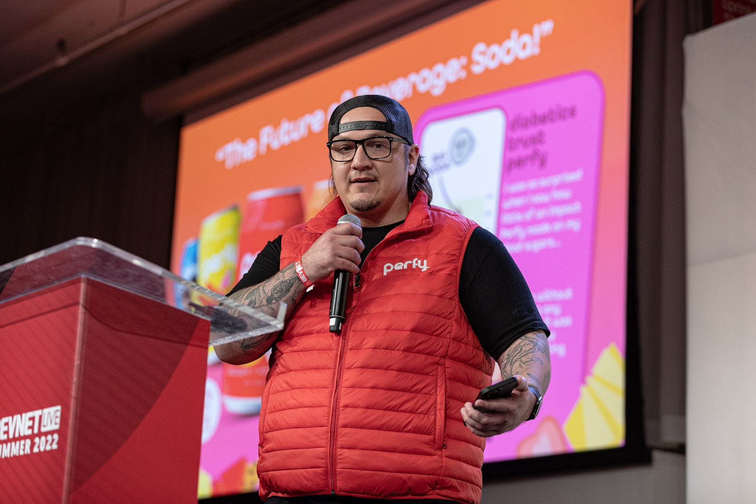 A man in a red Perfy puffer vest, glasses and a backwards cap holds a microphone and a phone in front of an orange slide.