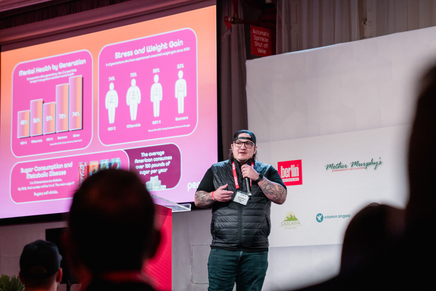 A man in a backwards cap, glasses and black puffer vest speaks into a microphone in front of a pink bar-chart slide, with audience silhouettes in the foreground.