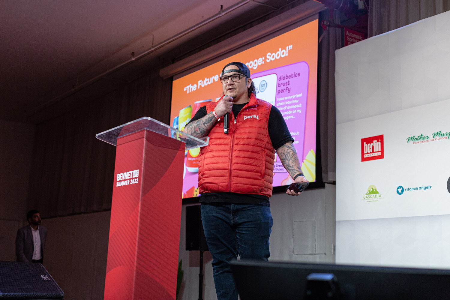 A man in a red vest holds a microphone at a red BevNET Live podium, a pink and orange slide behind him.