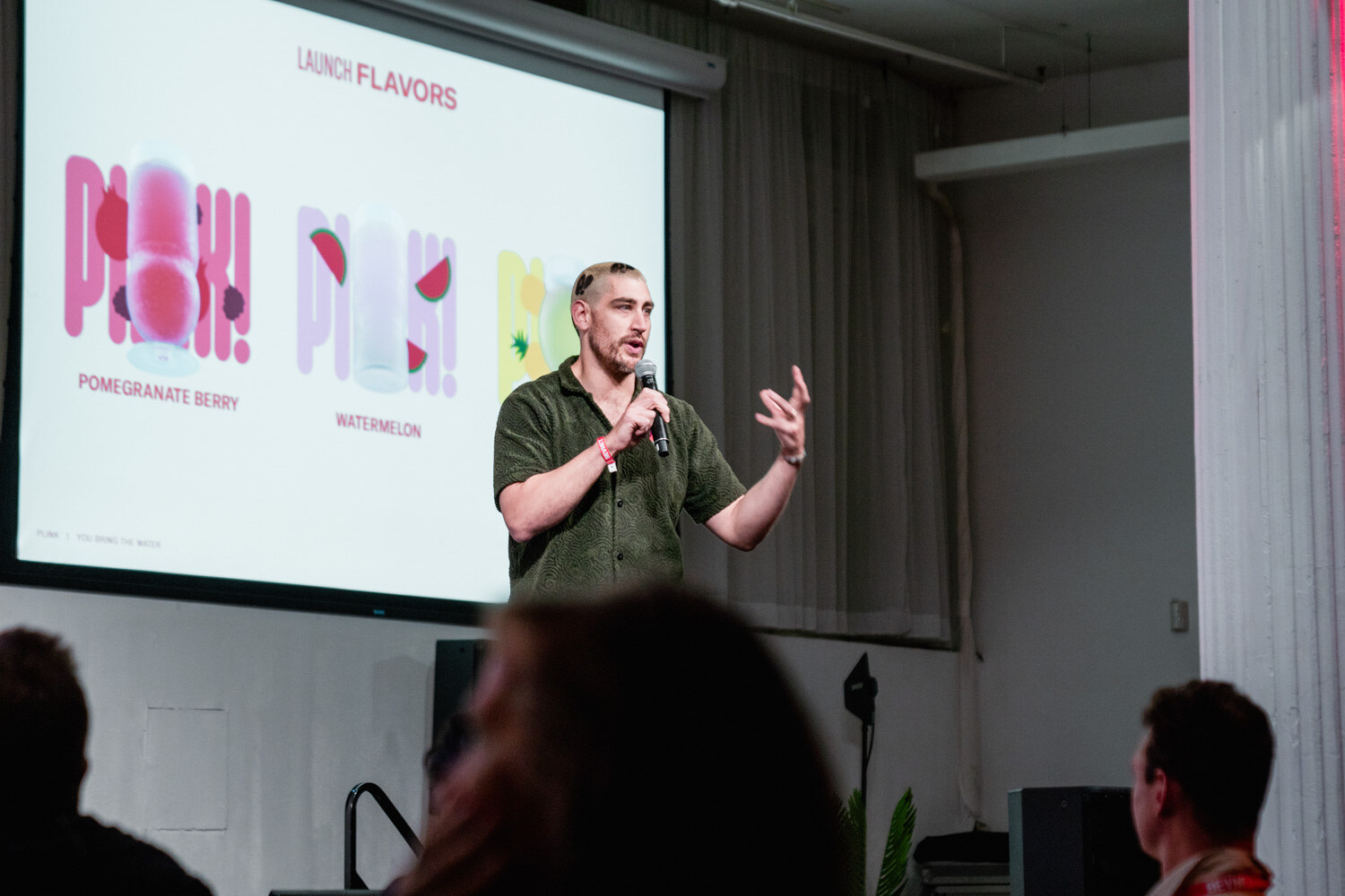 A man in a green shirt speaks into a microphone in front of a screen showing Plink launch flavors.