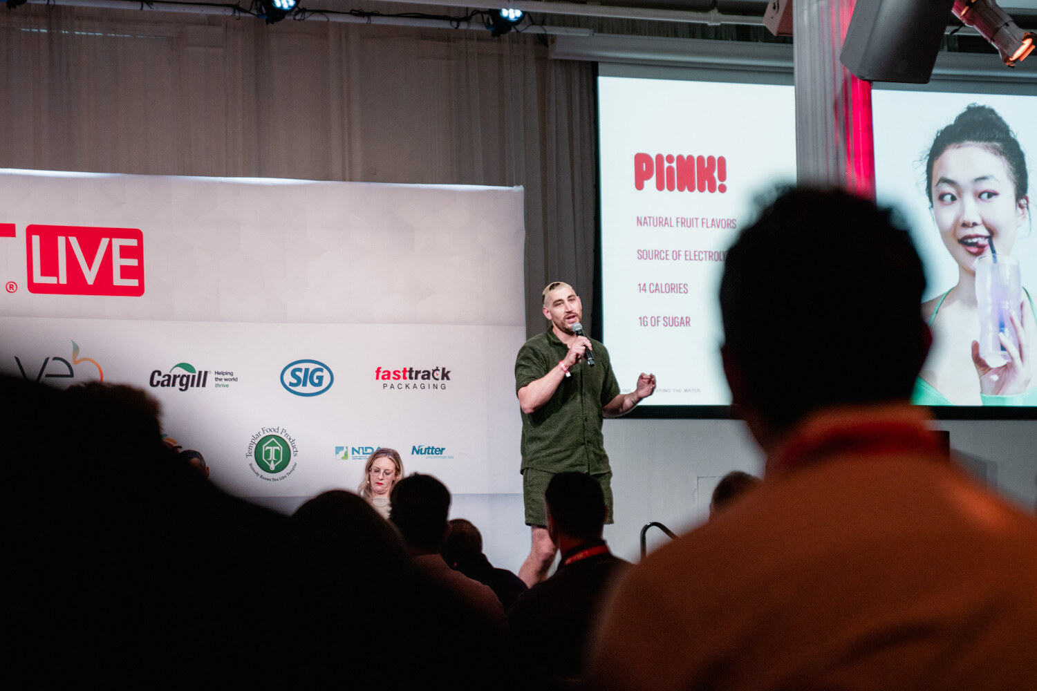 A man in a green shirt speaks into a microphone on stage as a screen shows a Plink slide with a woman drinking.