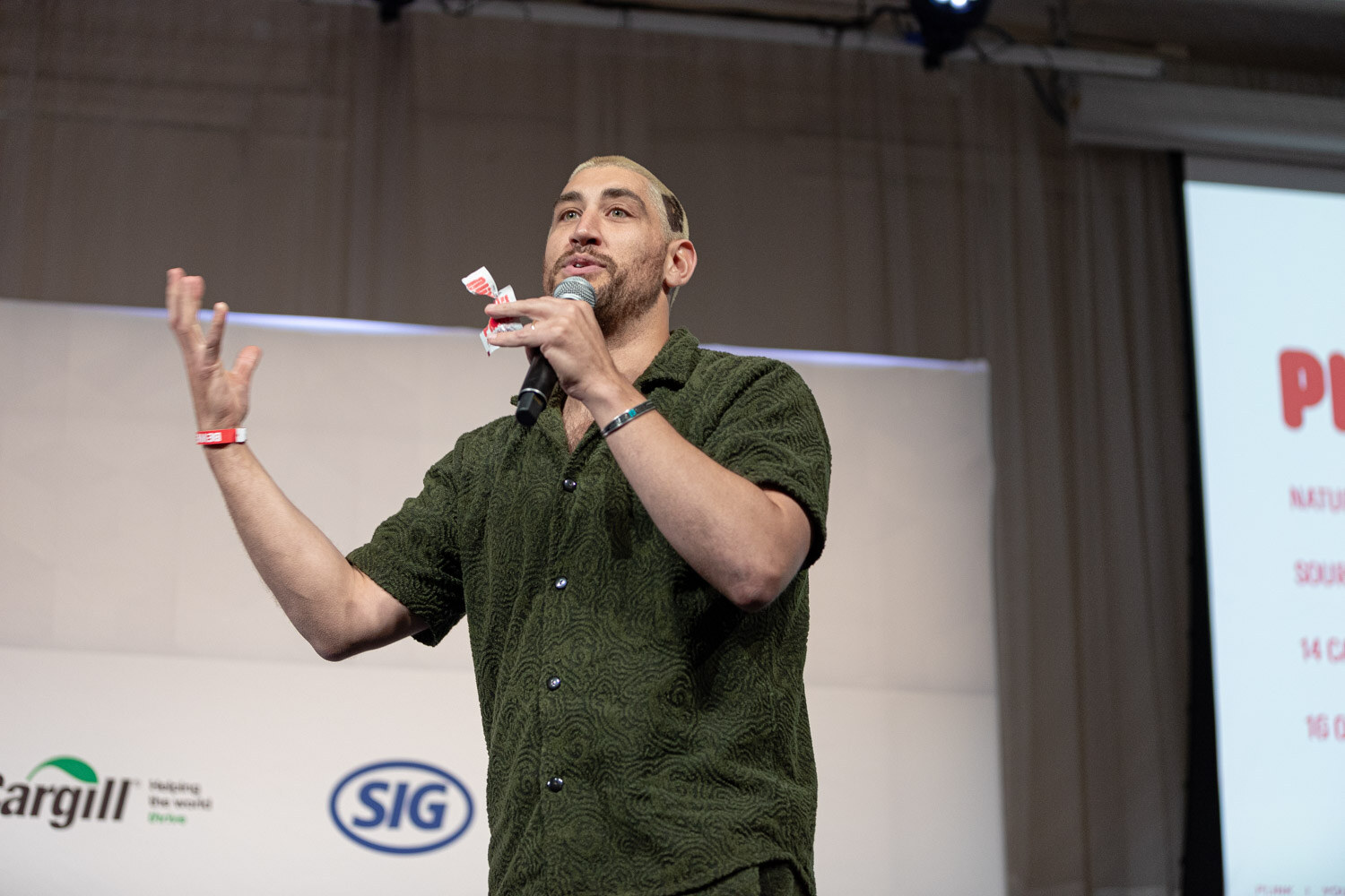 A man in a green shirt holds a microphone and a small paper and gestures dramatically on stage.