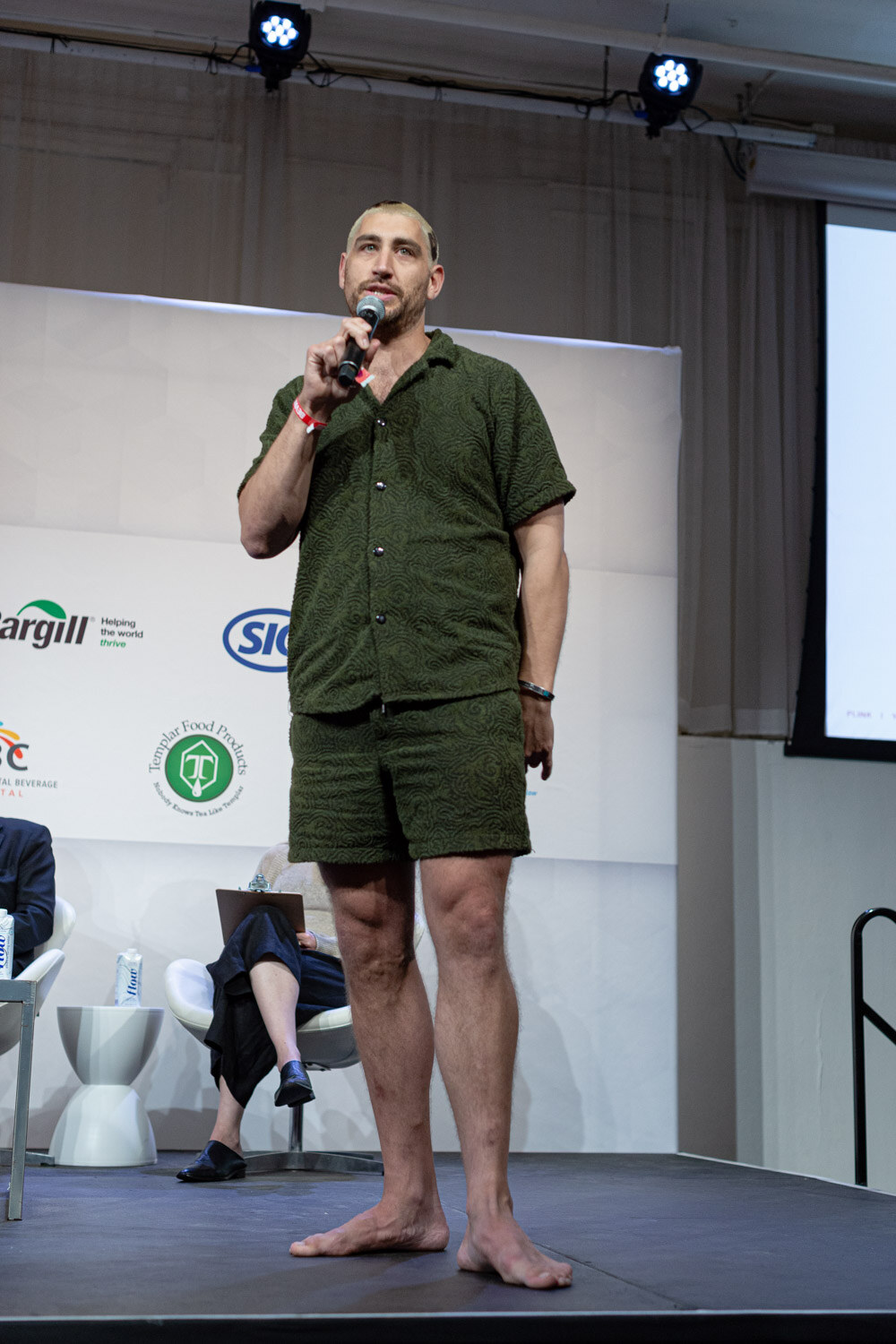 A man in a green short-sleeved shirt and matching shorts stands on stage with a microphone, a judge seated behind him.