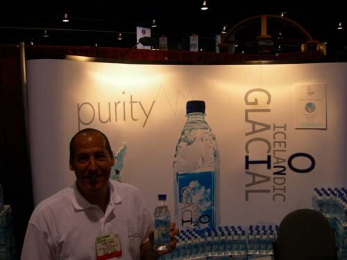 A smiling man in a white polo shirt holds a small water bottle in front of an Icelandic Glacial backdrop reading purity, with cases of bottled water stacked along the bottom.
