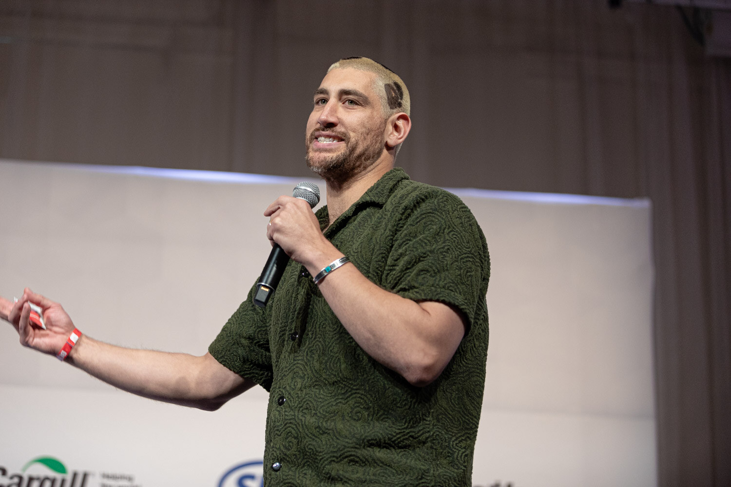 A man in a dark green short-sleeved shirt with a patterned haircut holds a microphone and gestures toward the audience.