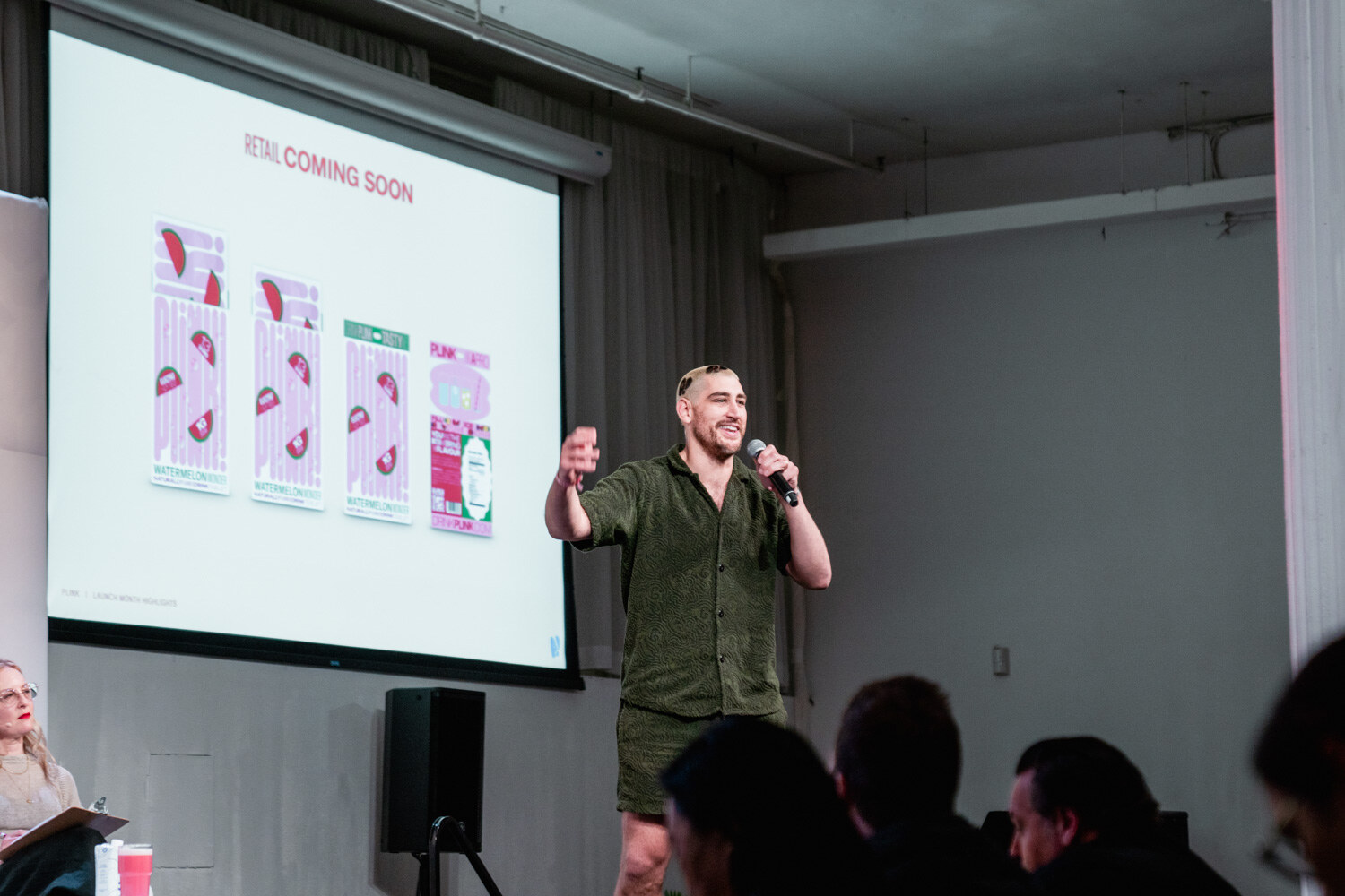 A man in a green shirt holds a microphone and raises one hand in front of a slide titled Retail Coming Soon.