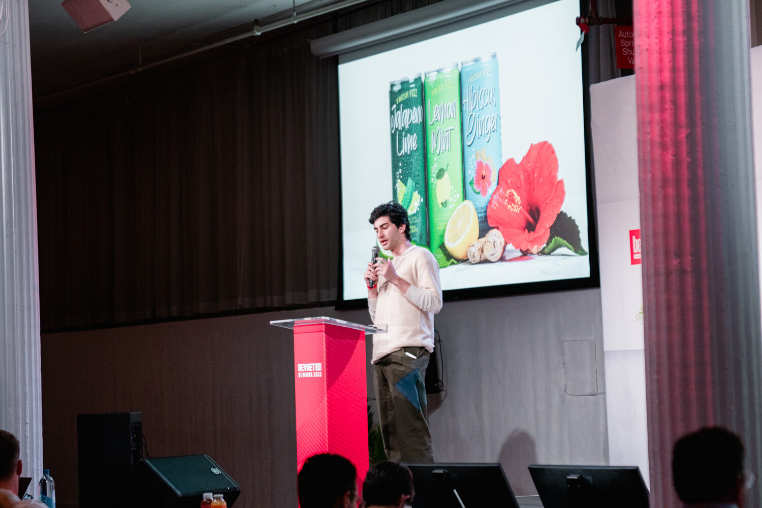 A man in a cream sweater stands at a red podium in front of a slide with three green cans, limes and a red flower.