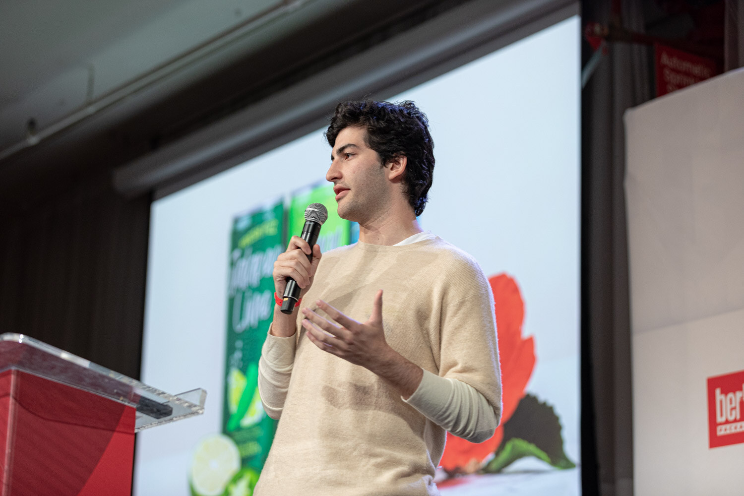 A young man in a cream sweater holds a microphone beside a red podium as a slide shows a green can with limes and a red flower.