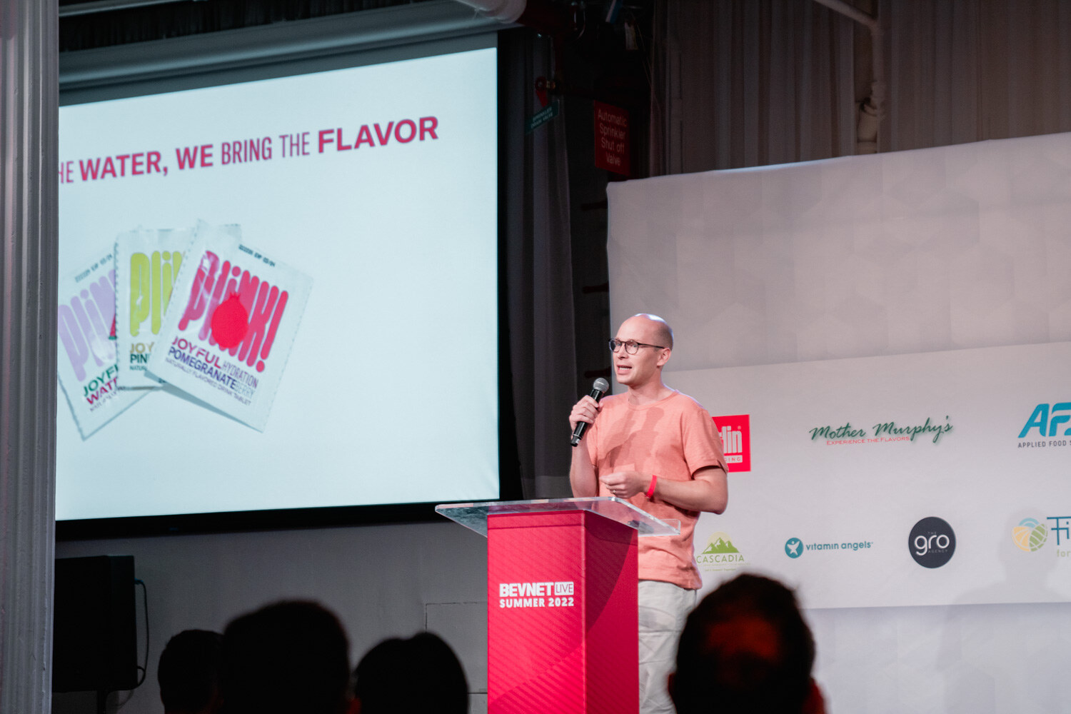 A bald man with glasses in a peach t-shirt holds a microphone at a red podium in front of a pink slide.