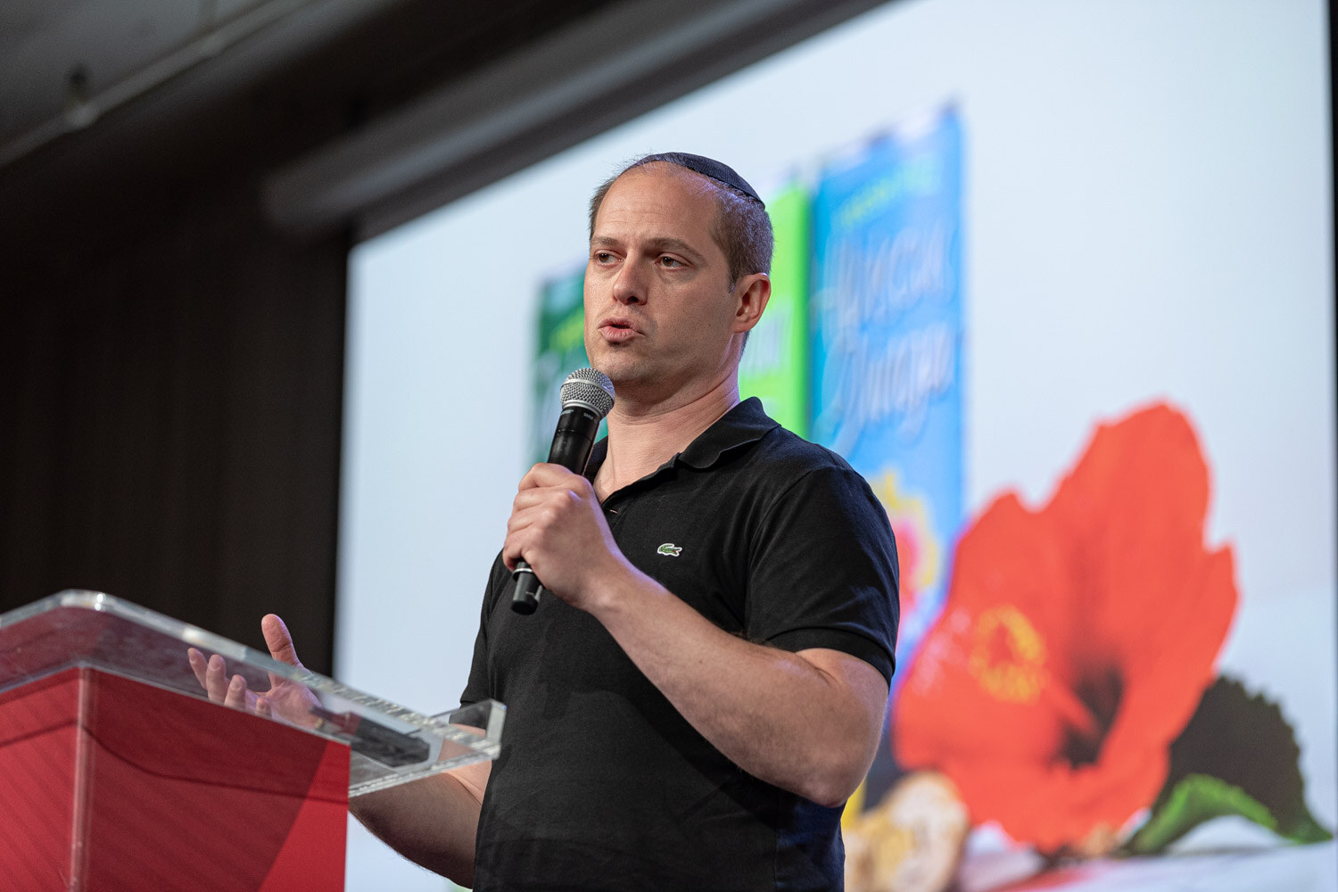 A man in a black polo holds a microphone at a podium with a slide showing a red hibiscus flower and a can behind him.