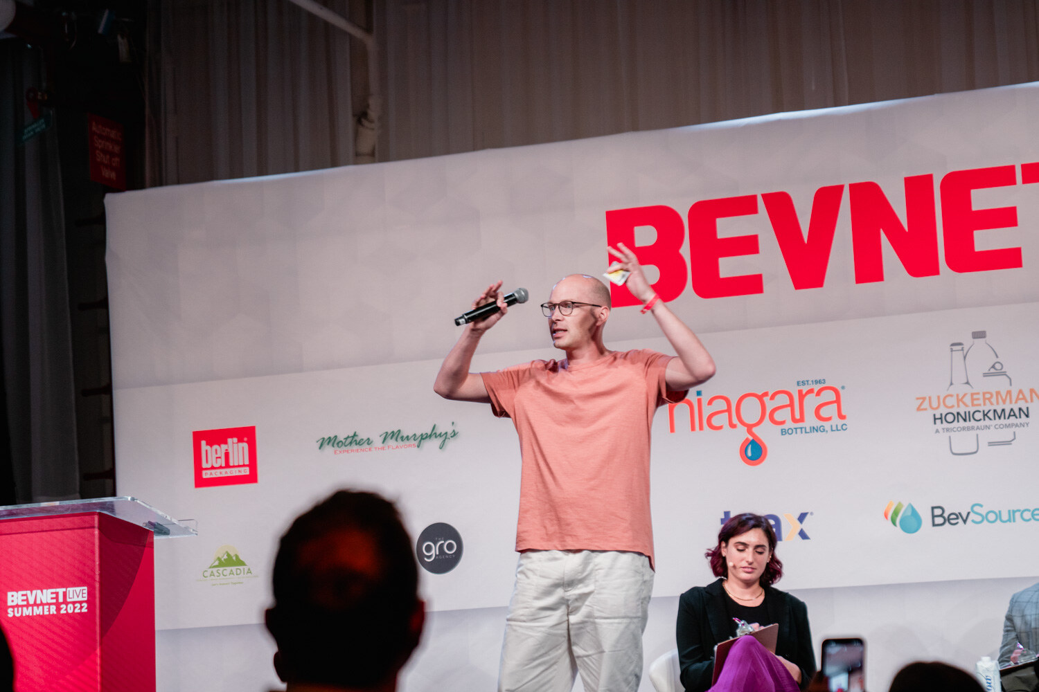 A bald man in a peach t-shirt holds a microphone and raises one hand on a stage below the BevNET Live sign.