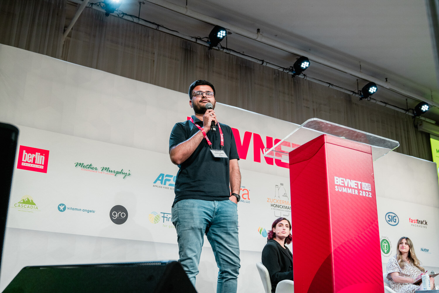 A man in a black polo and lanyard stands with a microphone in front of a BevNET Live backdrop, two judges seated in the background.