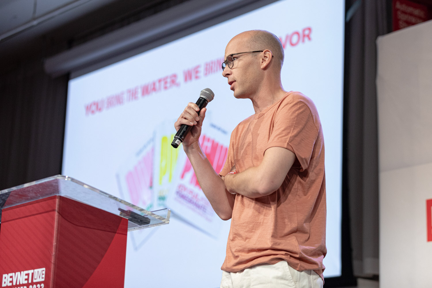 A bald man with glasses and a peach t-shirt holds a microphone with a hand across his chest, a slide behind him reading You bring the water, we bring the flavor.