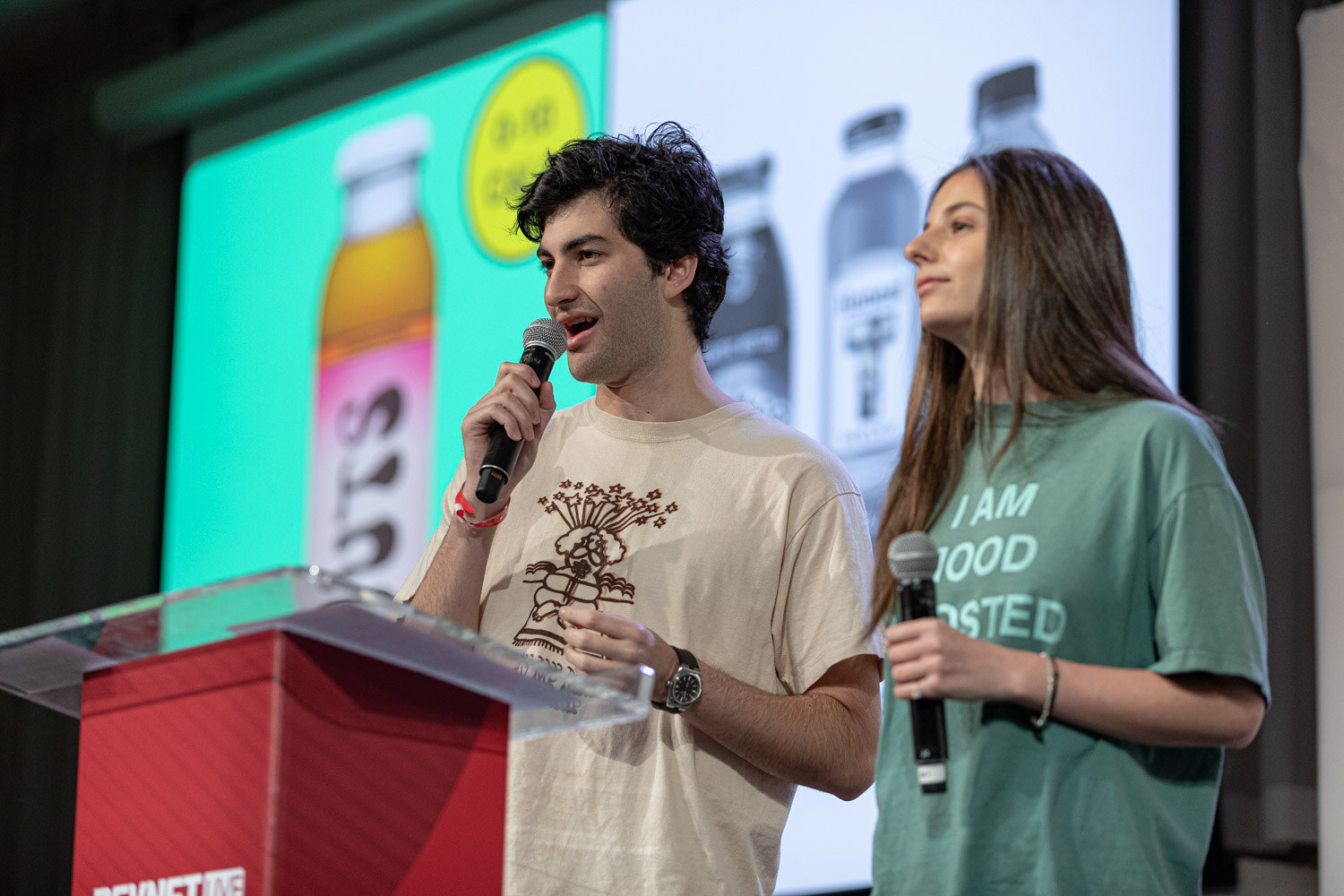 A man in a cream t-shirt and a woman in a green t-shirt each hold a microphone at a clear podium in front of a slide with bottled drinks.