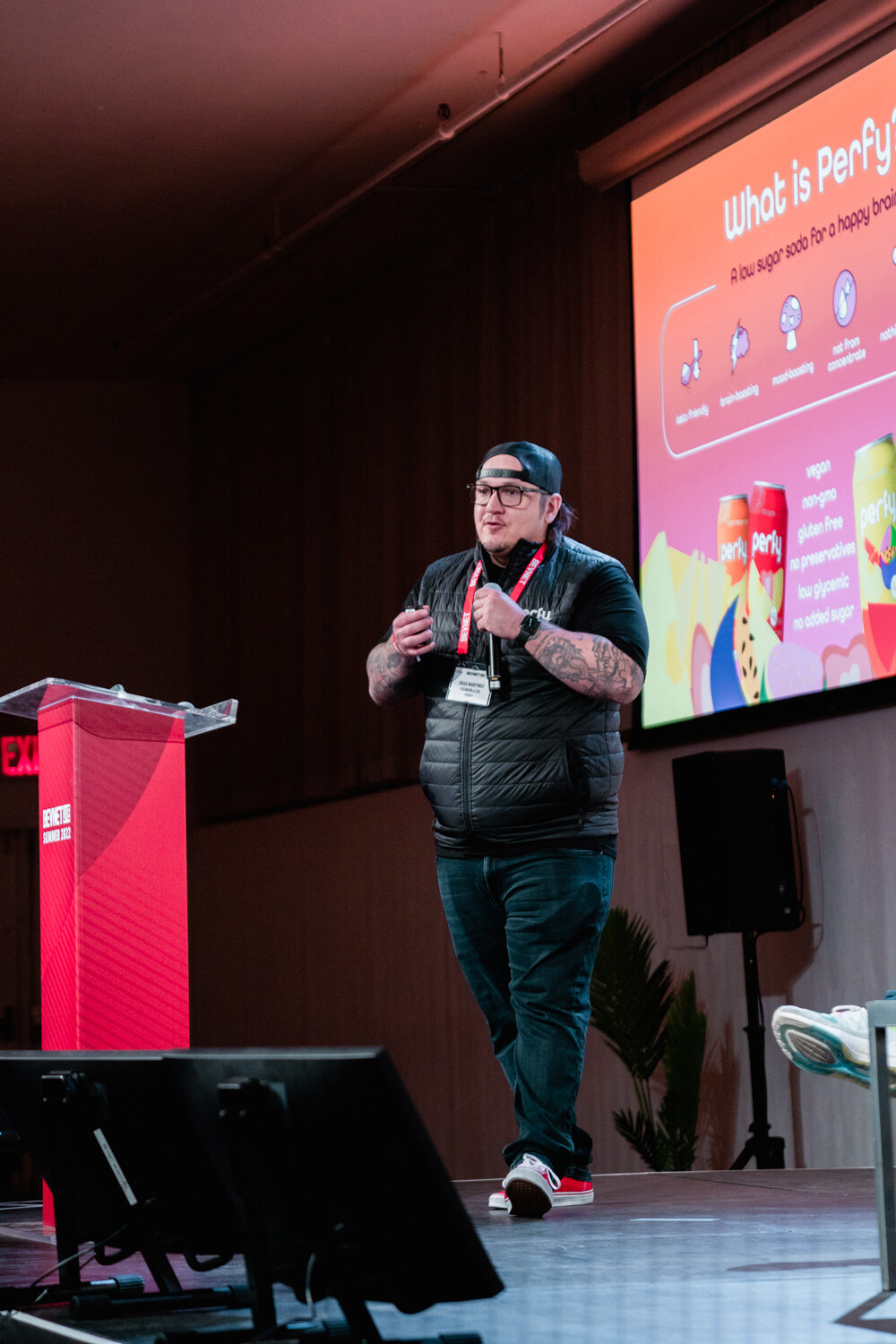 A man in a black vest and cap walks on stage with his hands together in front of a screen showing What is Perfy?