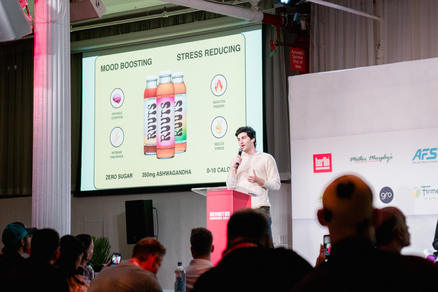 A man in a cream sweater stands by a red podium while a screen reads Mood Boosting, Stress Reducing with three Ruuts bottles.