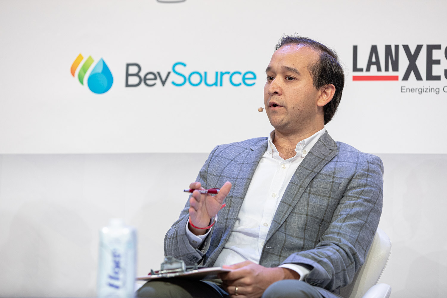 A man in a gray plaid blazer and white shirt gestures with a pen, seated in front of BevSource and Lanxess signs.
