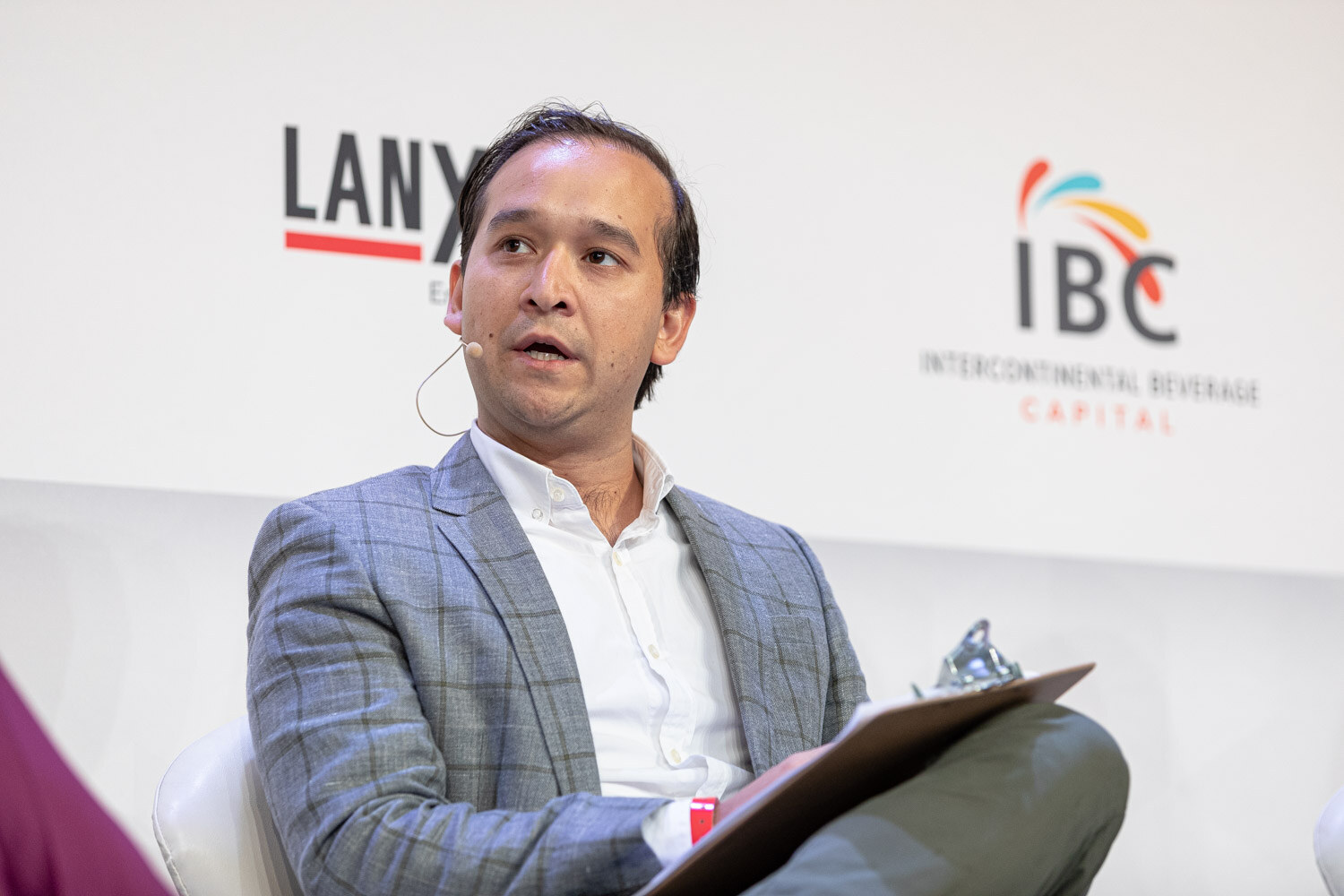 A man in a gray plaid blazer and white shirt speaks into a headset microphone with a clipboard in his lap, IBC and Lanxess signs behind him.