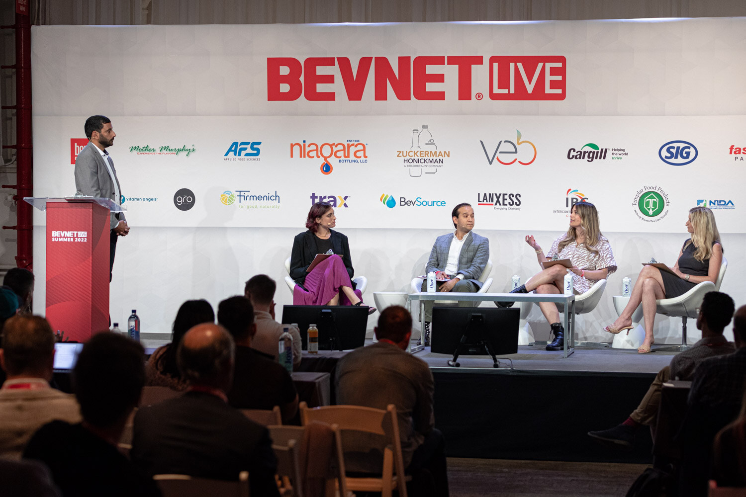 Wide view of a man at a podium and four seated judges on a stage below a BevNET Live backdrop, the audience's heads in the foreground.