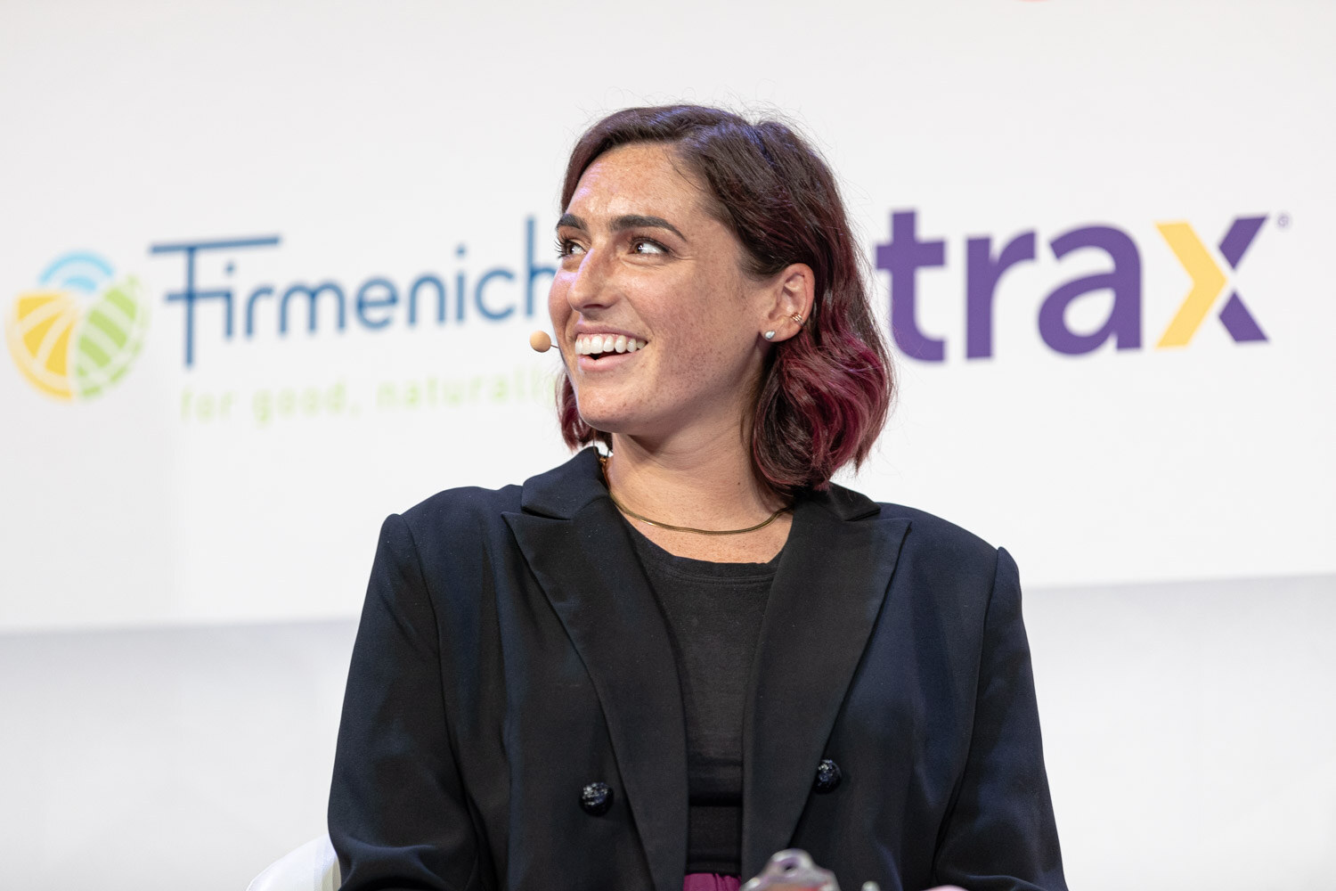 A woman with short wavy hair in a black blazer smiles in front of Firmenich and Trax signs.