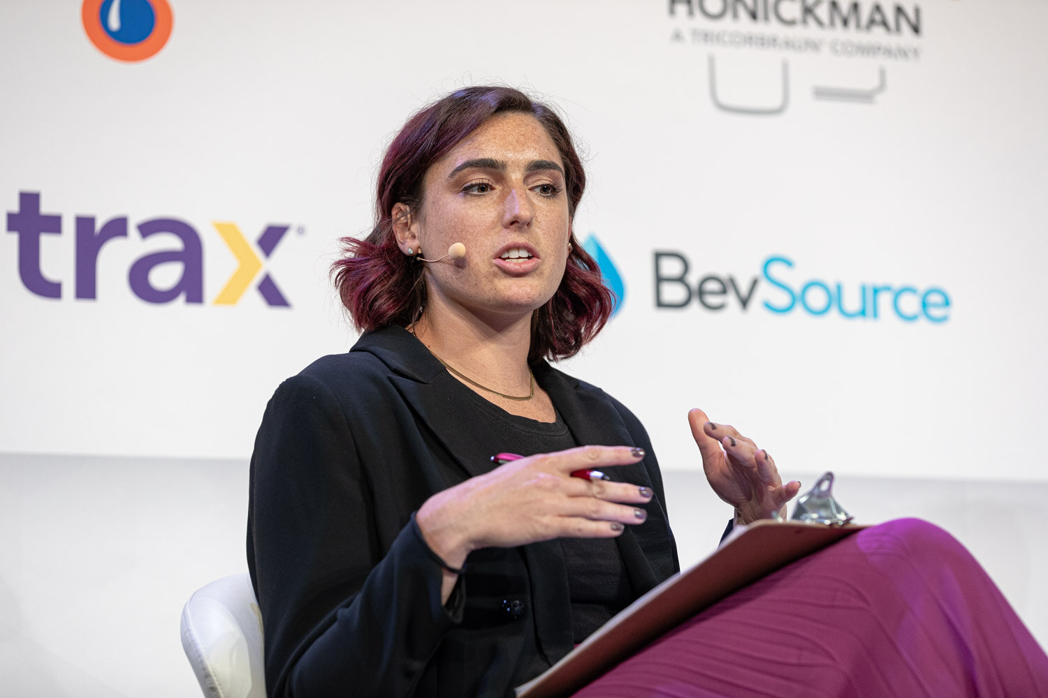 A woman with burgundy hair in a black blazer and magenta skirt gestures while speaking, with Trax and BevSource signs behind her.