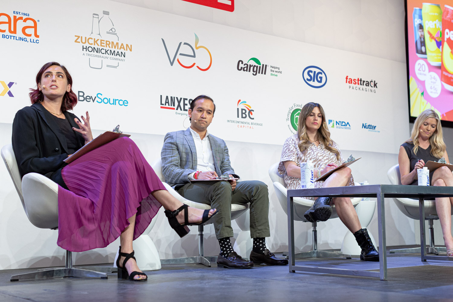 Four people seated in white chairs on a stage, a woman in a magenta skirt, a man in a gray blazer and two women, each with a clipboard.