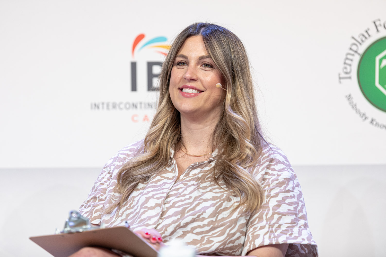 A woman with long blonde hair in a brown and white patterned dress smiles with a clipboard in her lap.