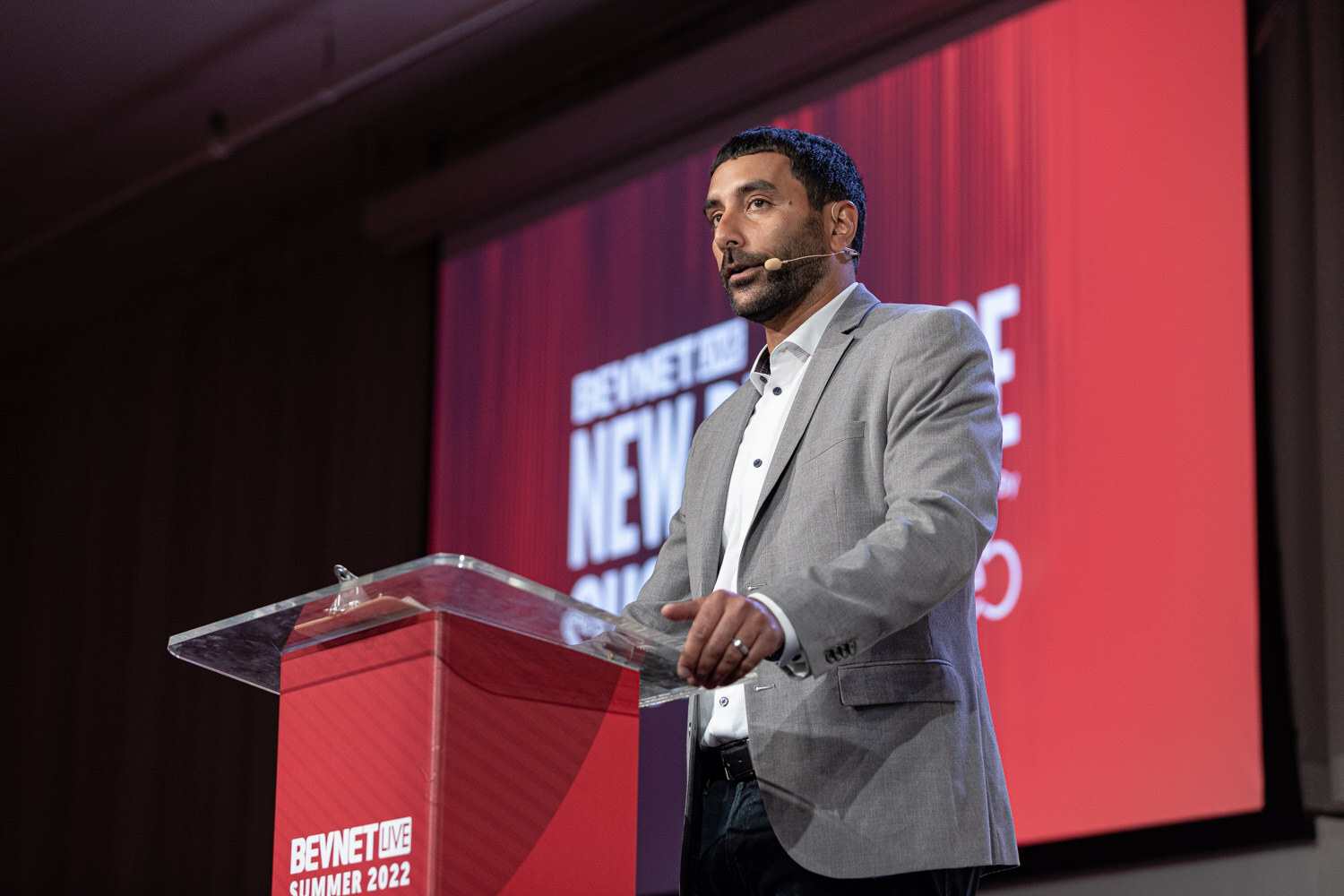 A man in a gray blazer and white shirt speaks behind a clear podium in front of a red screen reading New Beverage Showdown.