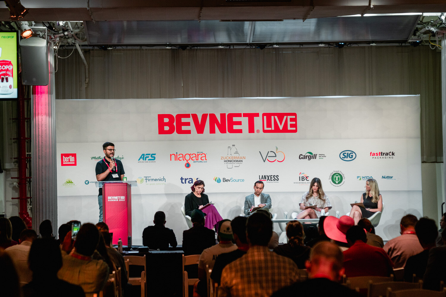 Wide view of a stage with a man at a red podium and four judges seated in white chairs under the BevNET Live sign.