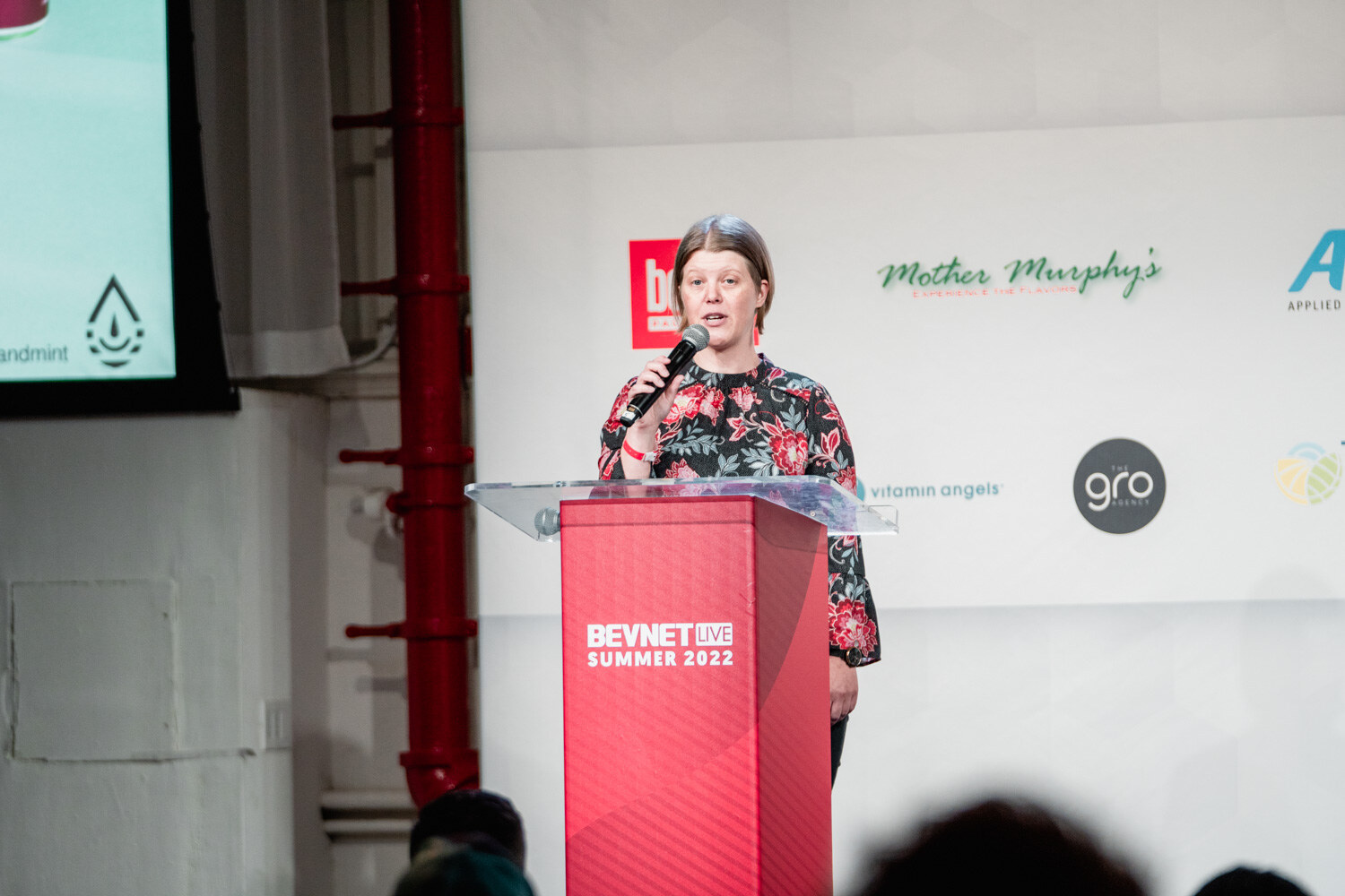 A woman in a floral blouse speaks into a microphone at a red BevNET Live podium with sponsor logos behind her.