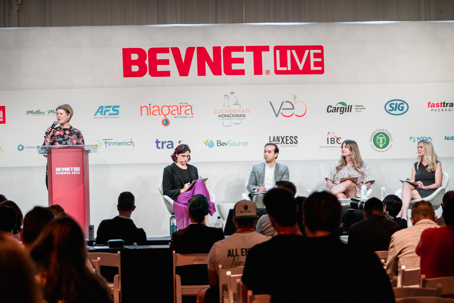 A woman in a floral top speaks at a red podium while four judges sit in chairs to her right in front of a BevNET Live backdrop.