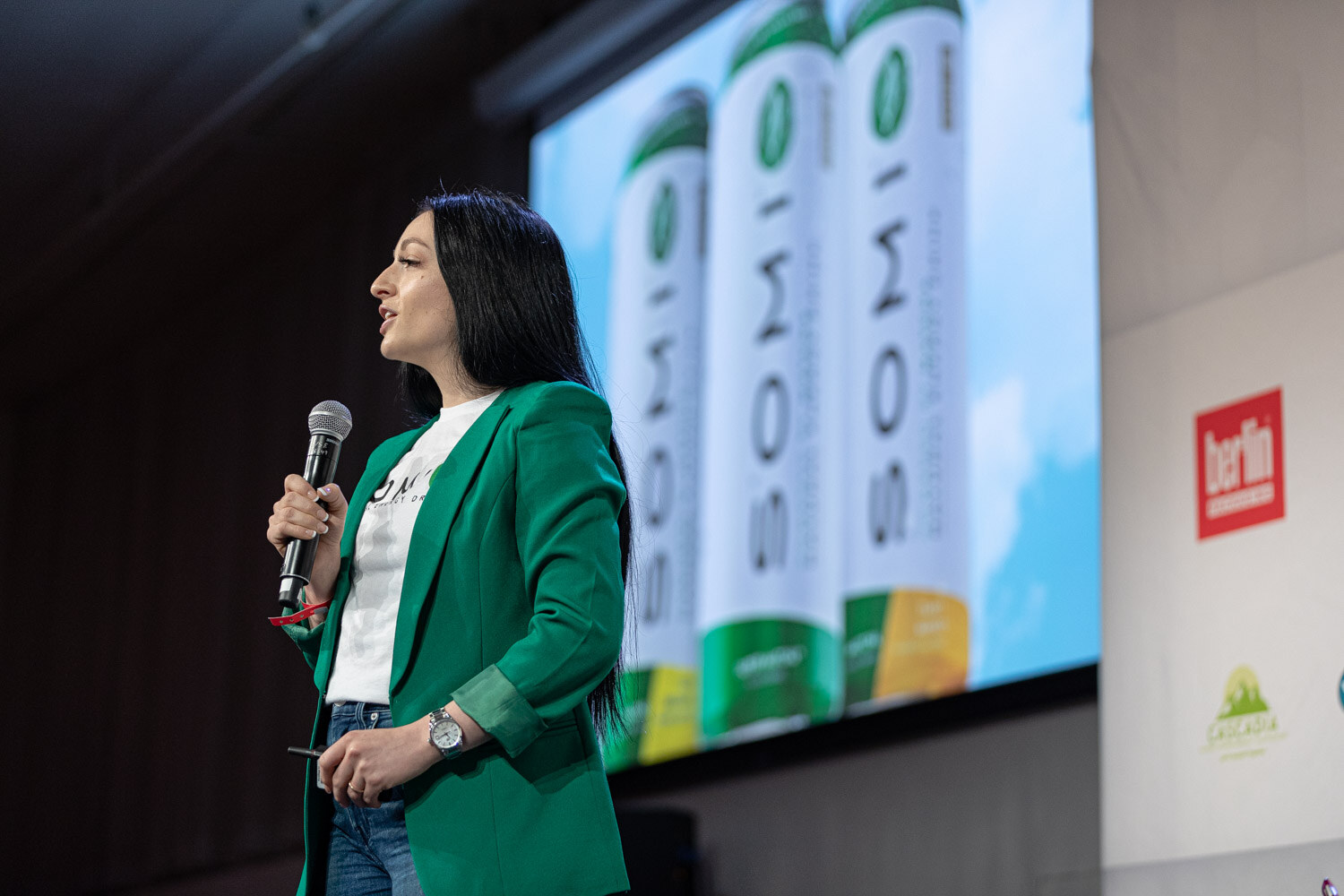 A woman in a green blazer holds a microphone and looks to her right in front of a screen showing three Somi cans.