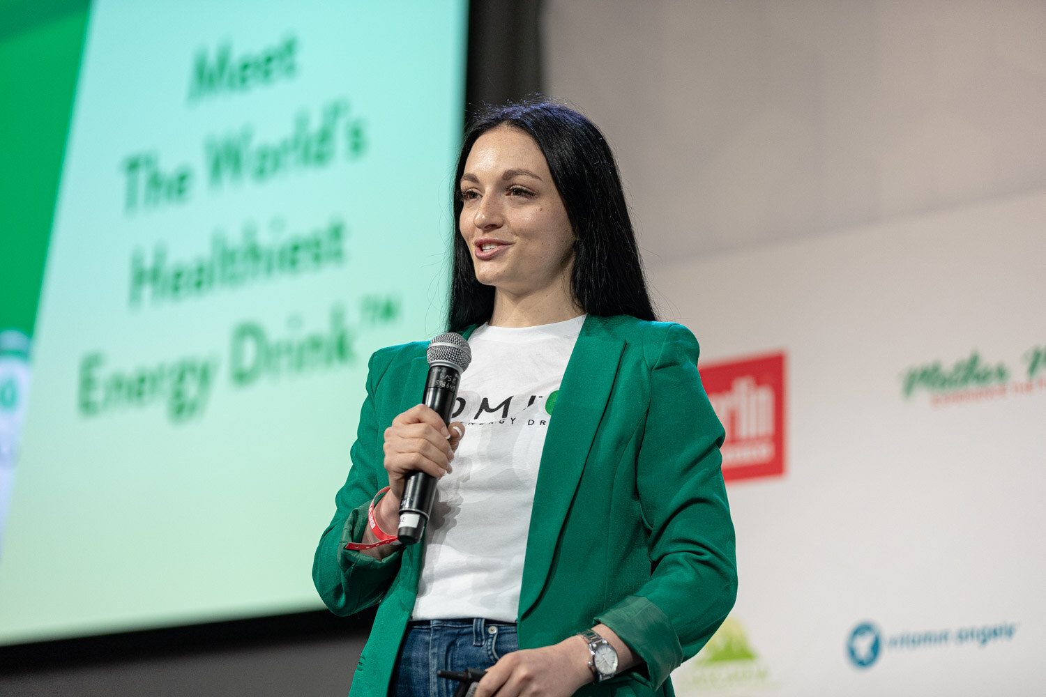 A woman with long dark hair in a green blazer and a white Somi shirt holds a microphone on stage, a slide behind her.