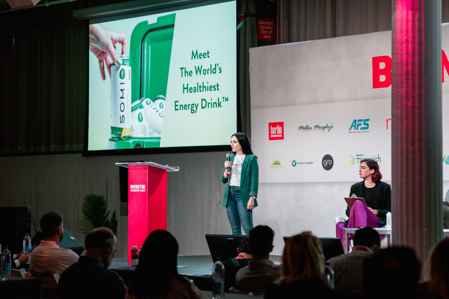 A woman in a green blazer stands on stage in front of a screen reading Meet the World's Healthiest Energy Drink, a seated judge at right.
