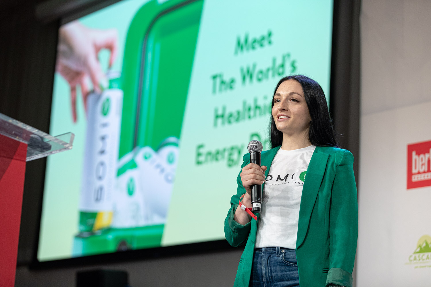 A woman in a green blazer holds a microphone in front of a mint green slide with a hand opening a Somi can.