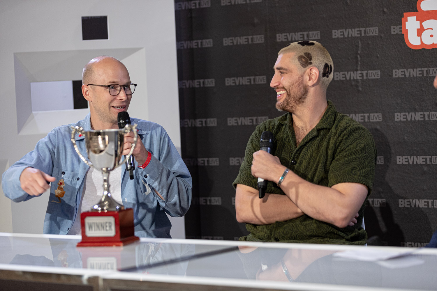 A bald man in glasses and a blue shirt holds a trophy and a microphone while a man in a green shirt laughs beside him.
