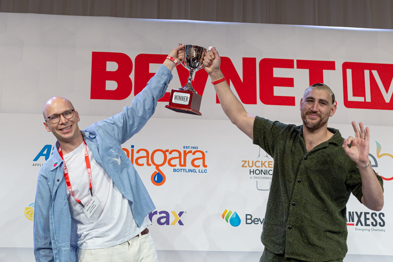 A bald man in glasses and a bearded man in a green shirt raise a trophy together under the BevNET Live sign, one making an OK sign.