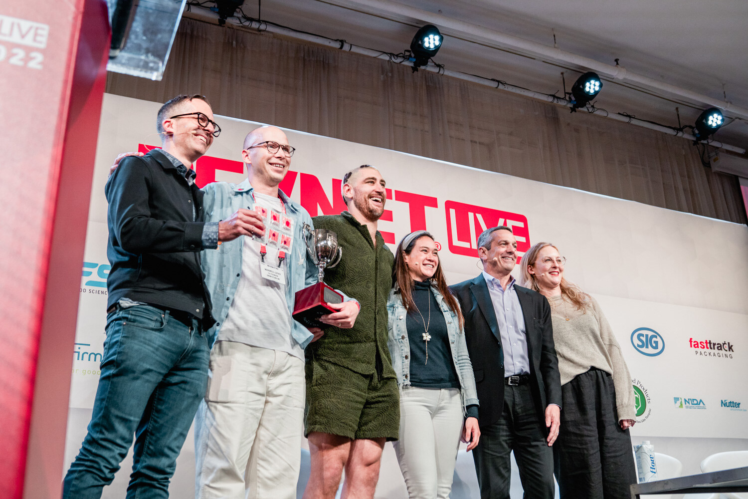 Seven people stand in a row on a stage below a BevNET Live sign, two men in the center holding a small trophy.