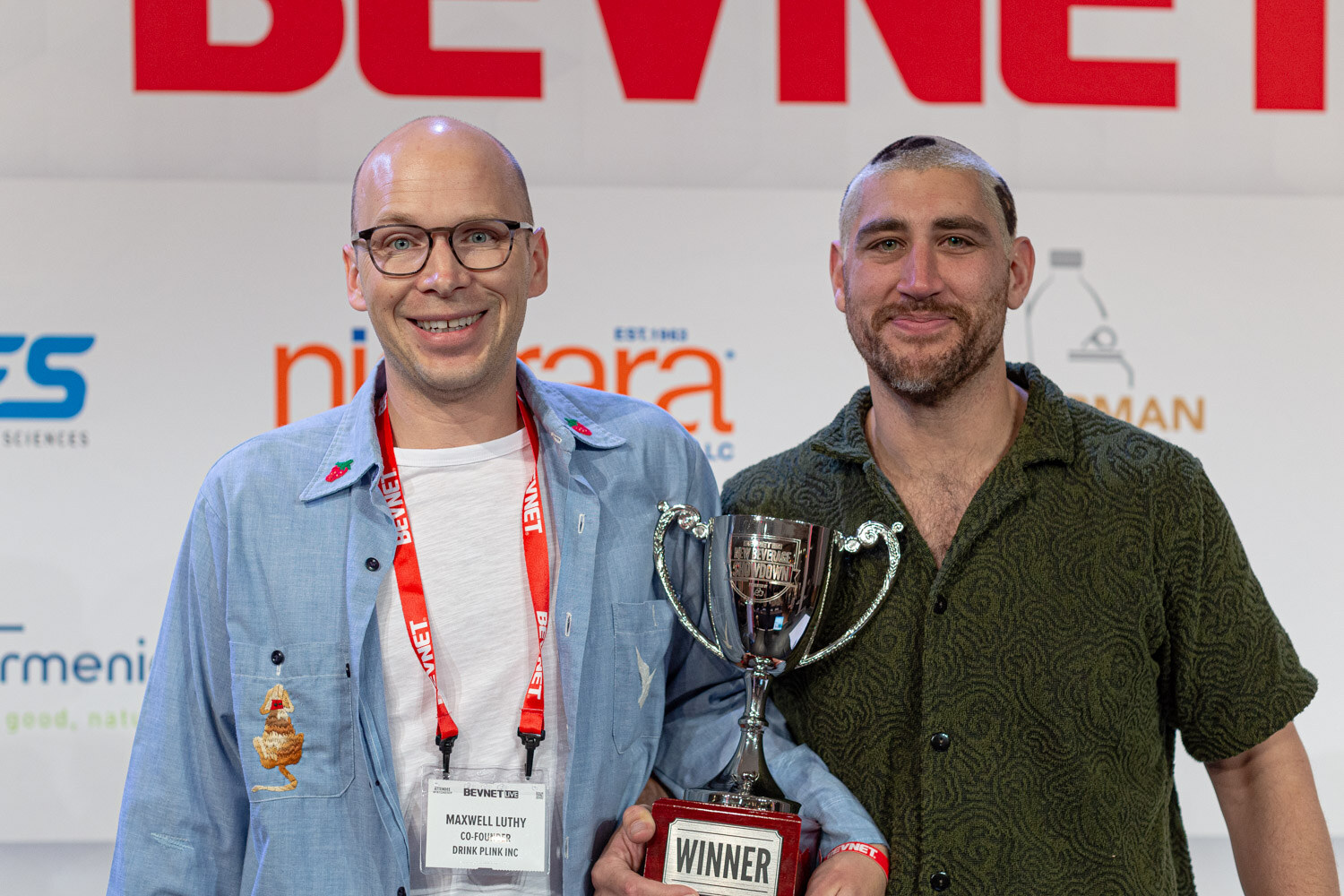 A bald man in glasses with a BevNET lanyard and a bearded man in a green shirt hold a silver winner's cup in front of a sponsor wall.
